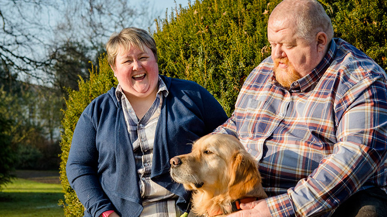 Two people sitting on a park bench with a guide dog and smiling